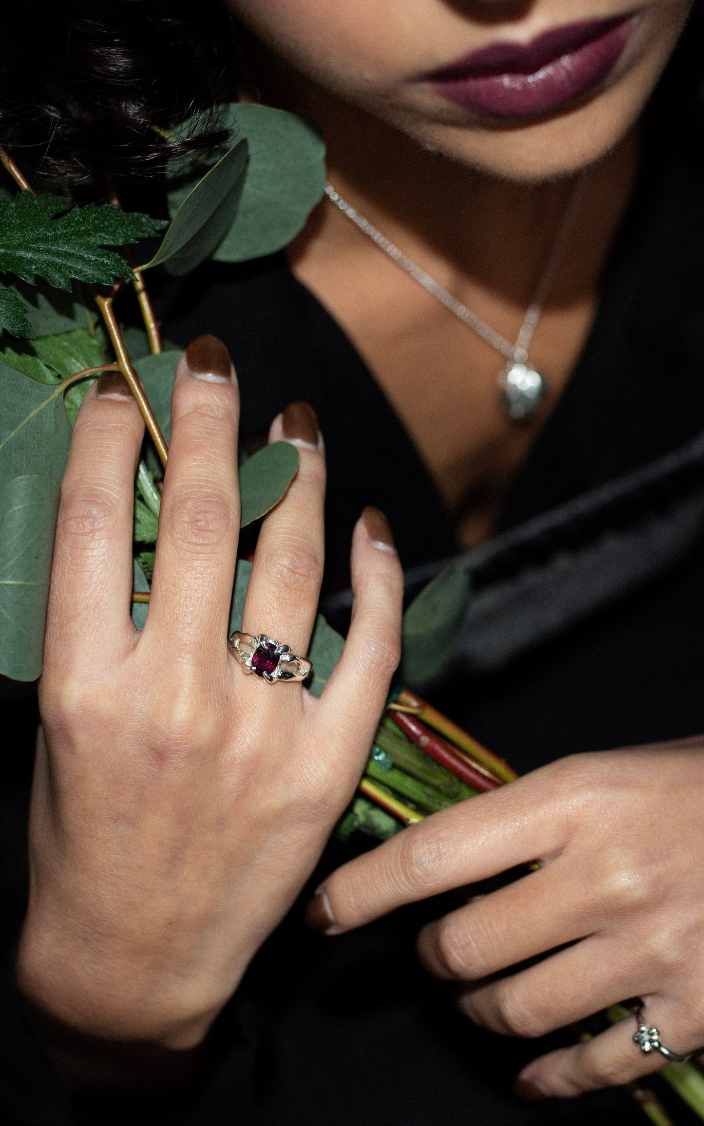 Close-up of a hand wearing a ring with a purple burgundygemstone, holding green leaves.