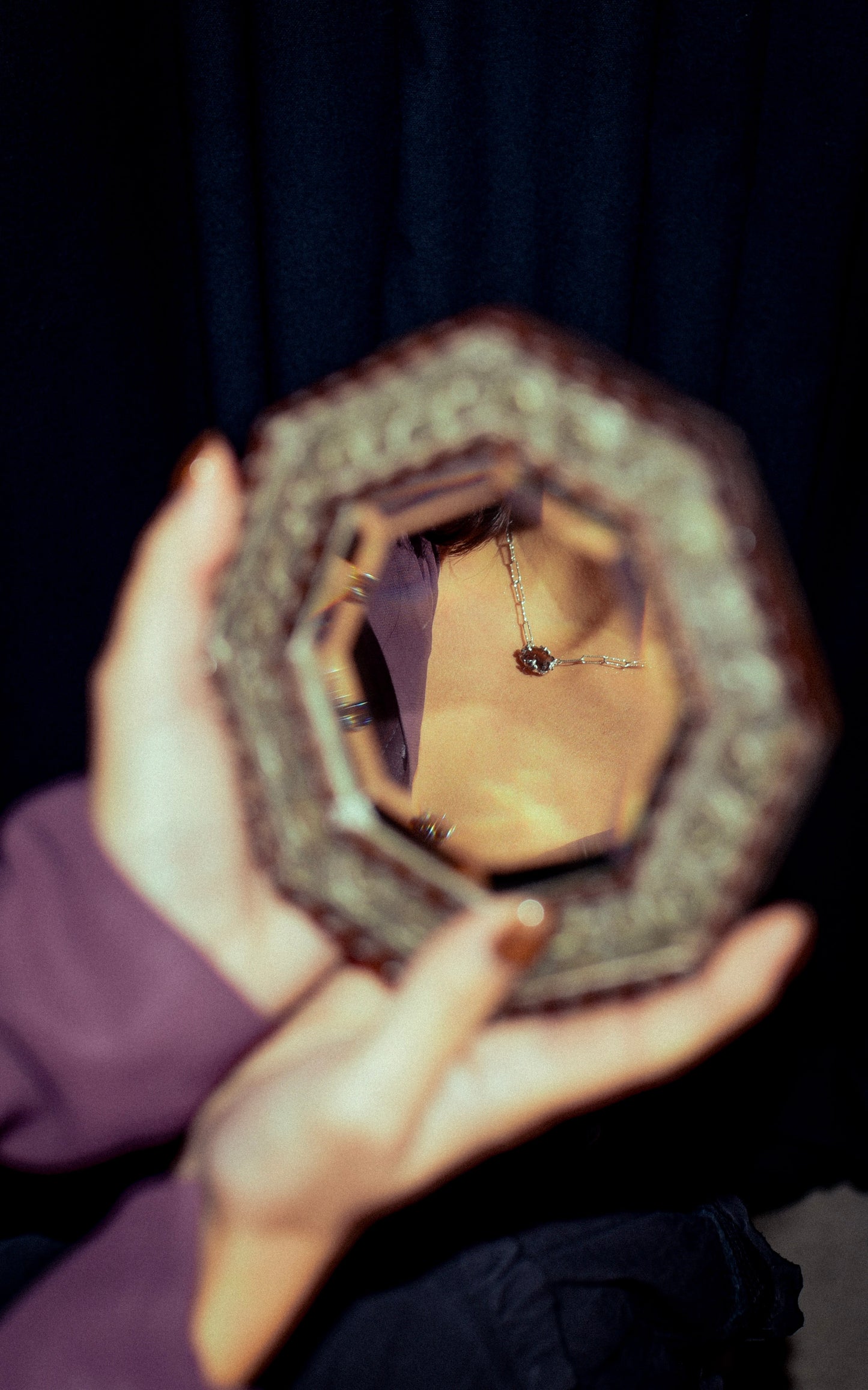 Person wearing a silver necklace, holding a hexagonal mirror with a dark background