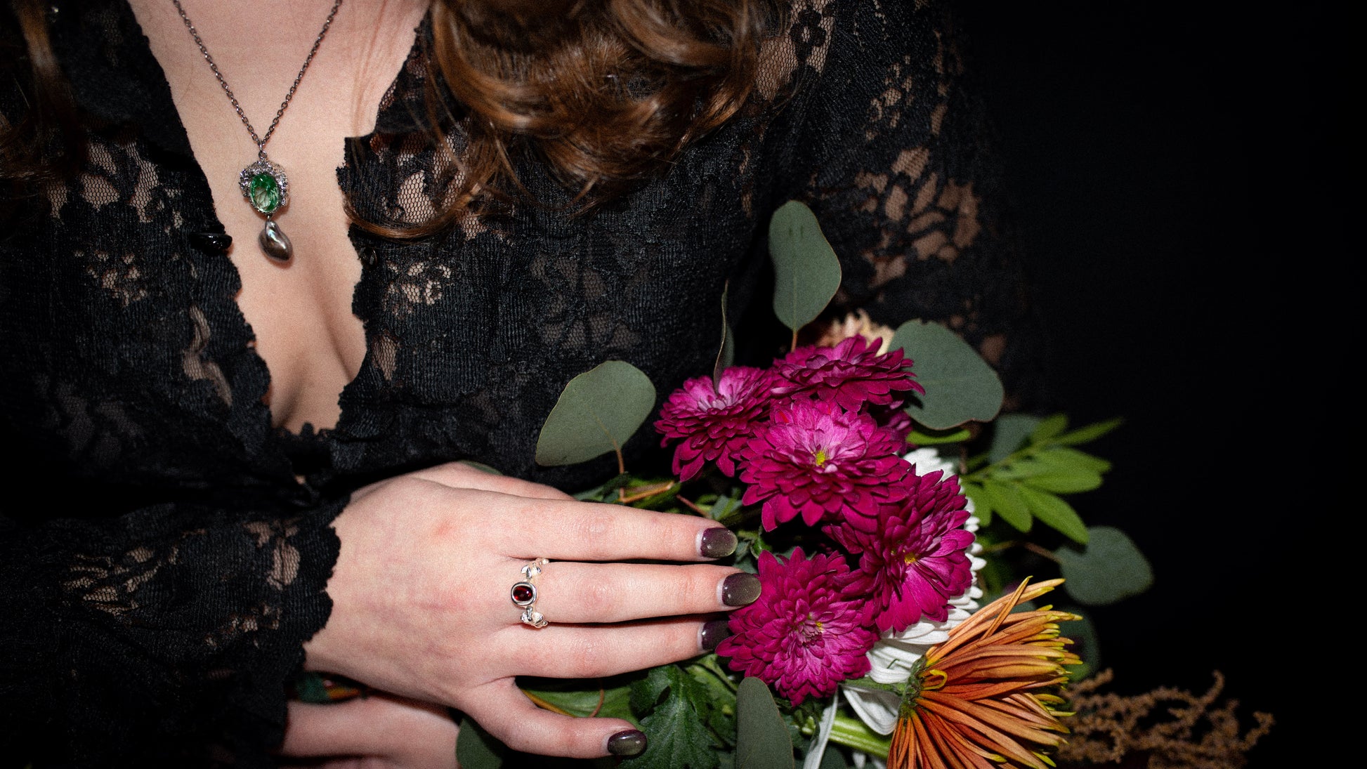 Person holding a bouquet of flowers with a dark background, while wearing a silver necklace with green gemstone and a silver ring with red gemstone