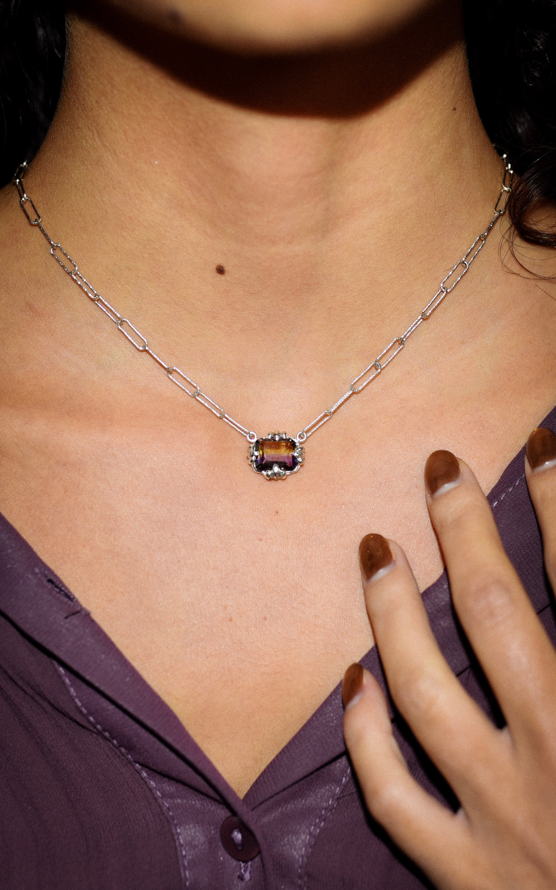 Silver necklace with a central purple and yellow gemstone worn by a person, with a hand touching the neckline.