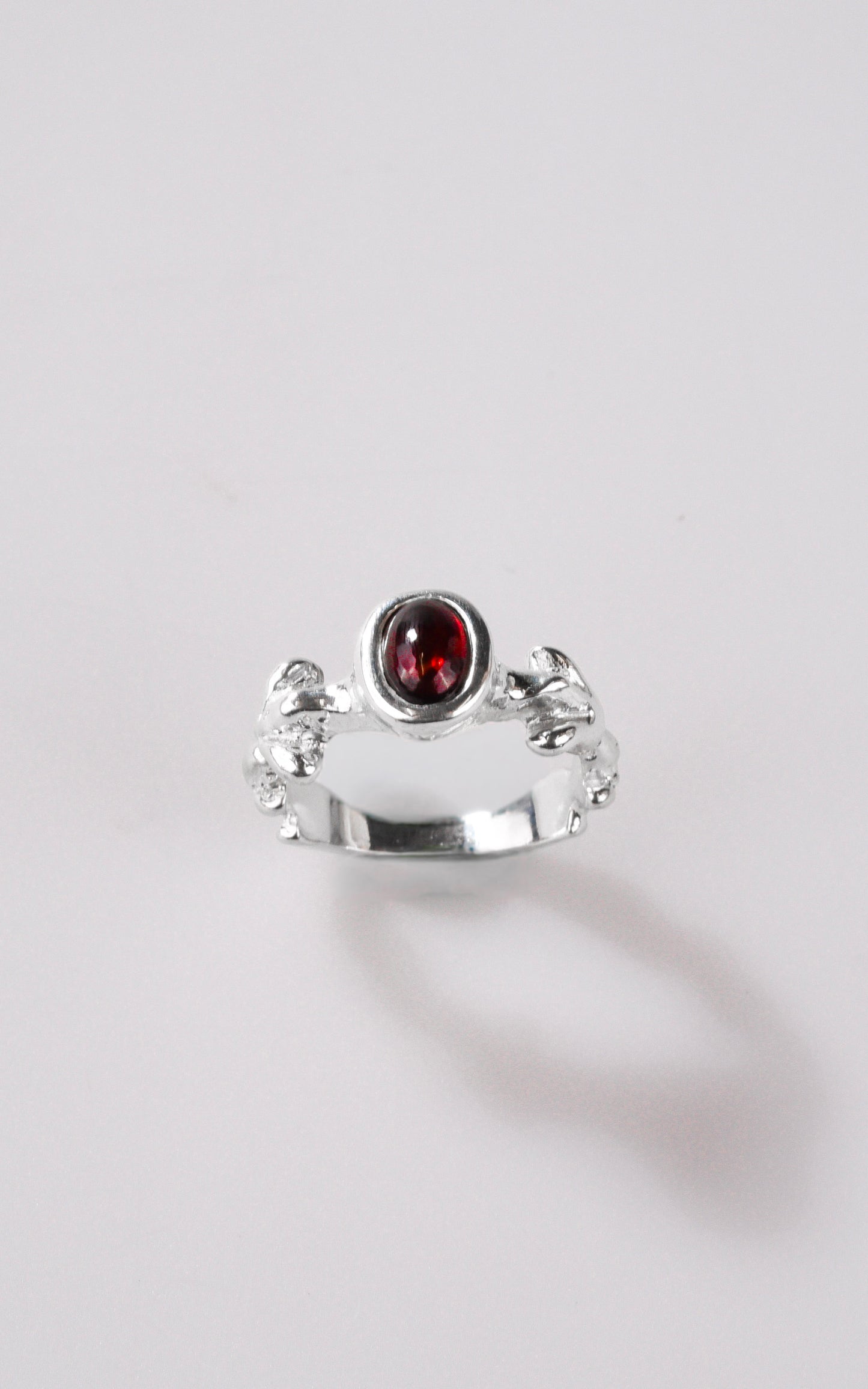 Silver ring with dark red oval gemstone and organic details on ring shank, with a light grey background