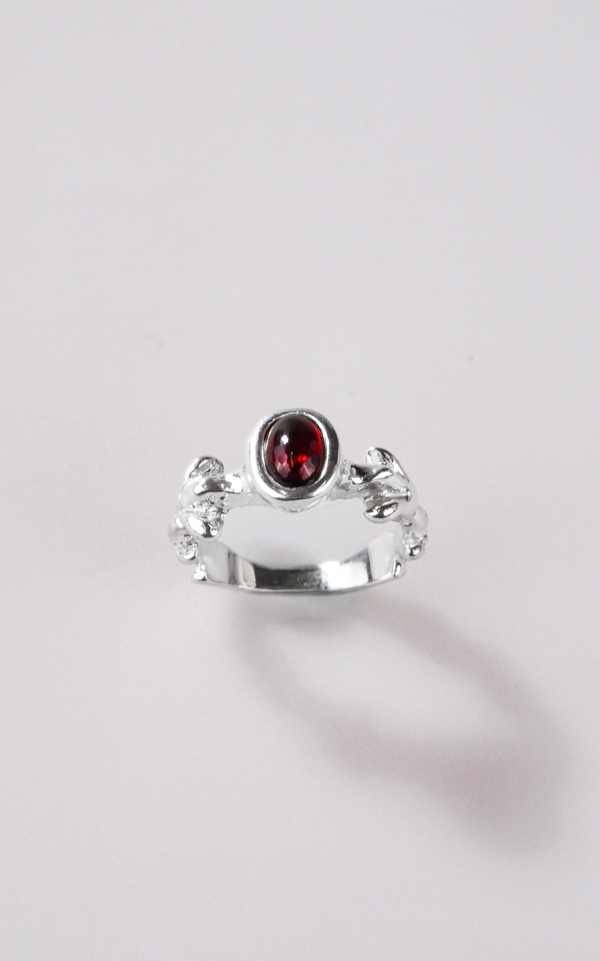 Silver ring with dark red oval gemstone and organic details on ring shank, with a light grey background