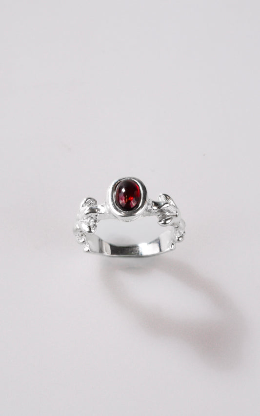 Silver ring with dark red oval gemstone and organic details on ring shank, with a light grey background