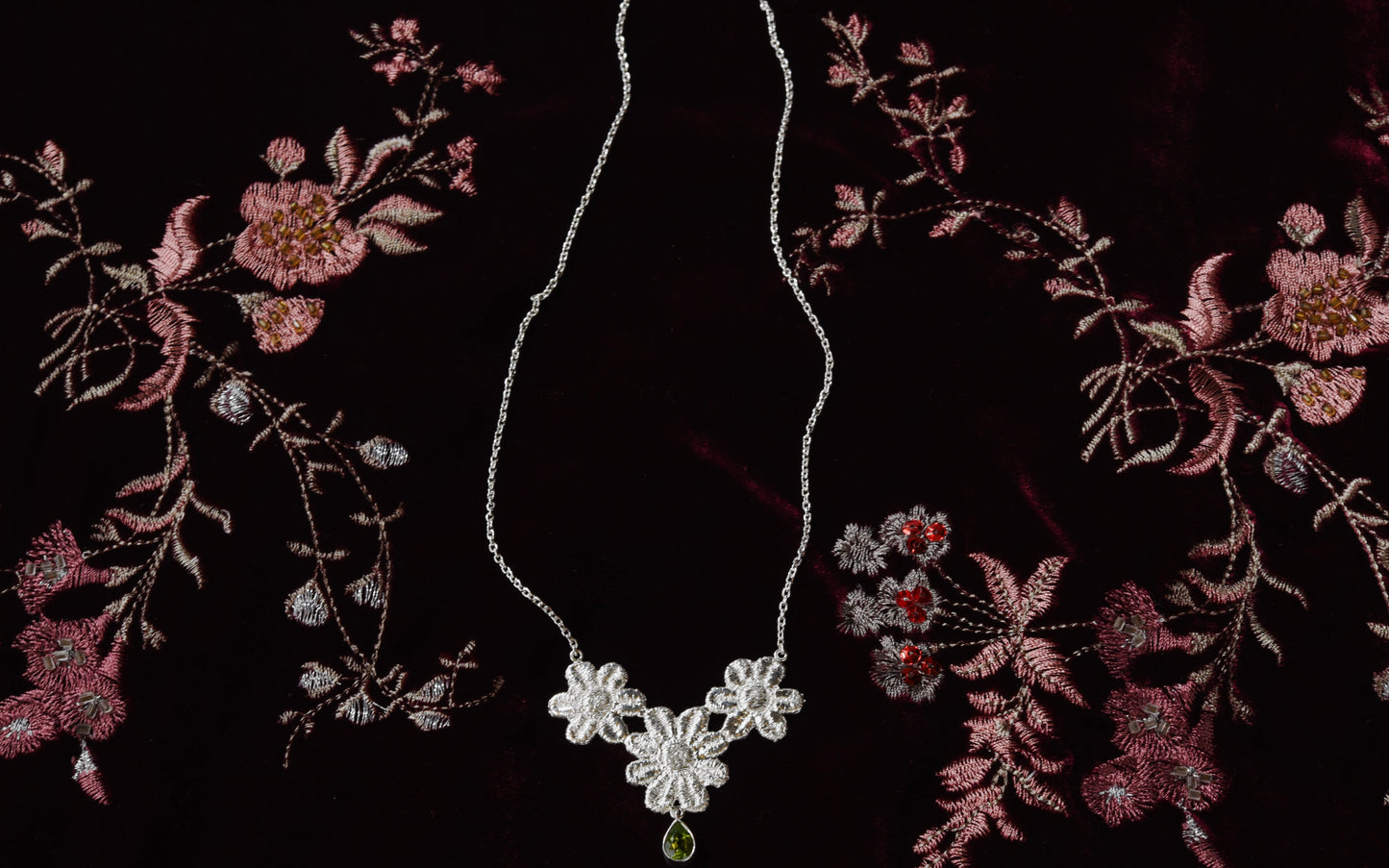 Silver necklace with floral lace pendant and green gemstone on a dark red velvet fabric background with floral embroidery