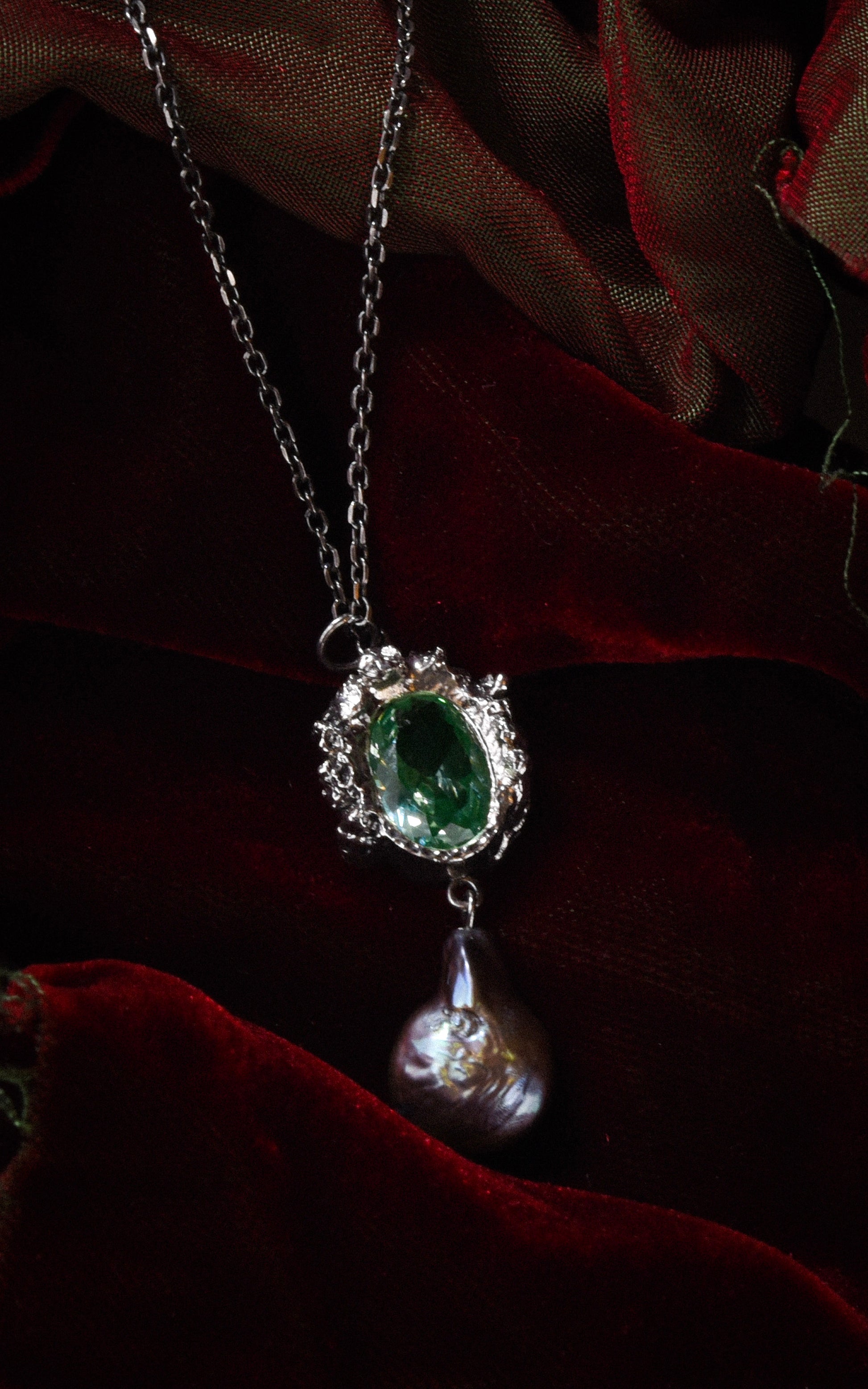 Silver necklace with a green gemstone pendant and purple baroque pearl on a dark red velvet background